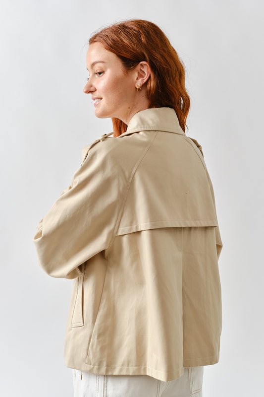 Back view of a woman wearing a beige cropped trench coat with storm flap detail and belted sleeves