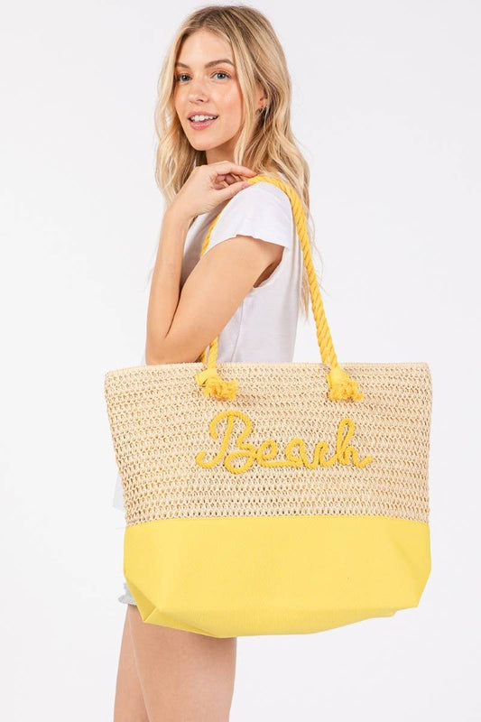 Blonde woman wearing a white tee and holding a large yellow and natural straw beach tote with "Beach" embroidered in yellow rope.