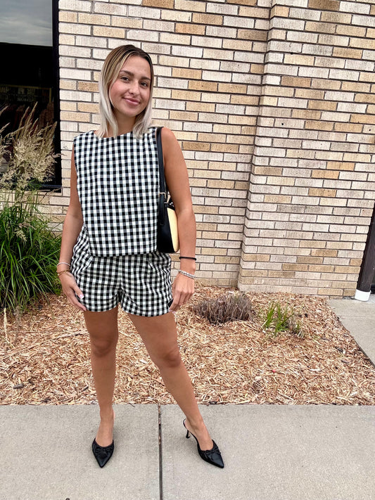 Woman wearing a black and white gingham matching set with a sleeveless boxy top and high-waisted shorts, styled with black heels and a structured handbag.