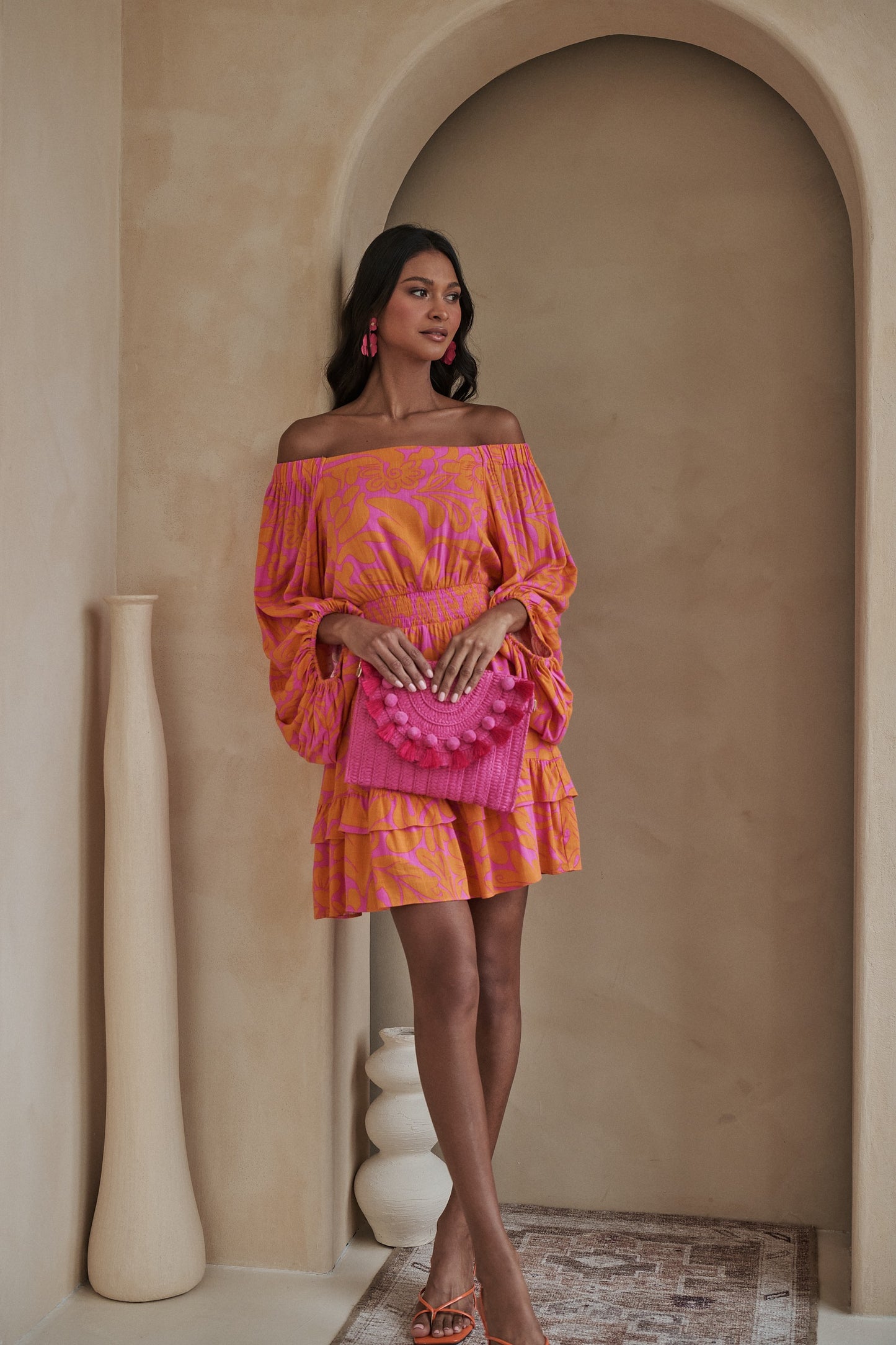 Woman wearing a pink and orange off-the-shoulder smocked mini dress with tiered ruffle hem now at Erin Rose Boutique.