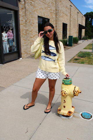 Woman in a yellow sweater and polka dot shorts standing next to a yellow fire hydrant on a sidewalk.