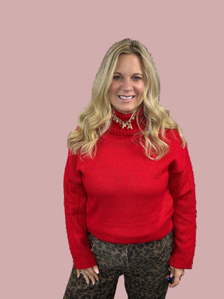 A woman standing, smiling, against a heart-patterned background. She is wearing a vibrant red turtleneck sweater with pearl details and leopard print pants.