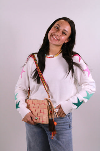 Woman wearing a white sweater with colorful star patterns, holding a woven handbag against a plain background