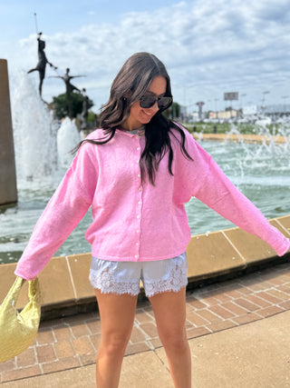 Woman wearing a pale pink button up cardigan sweater with lace shorts & butter yellow woven handbag.  