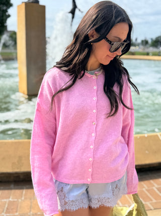 Woman wearing a pink cardigan and sunglasses standing near a fountain.