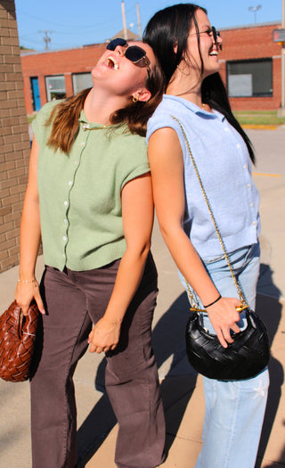 Two women posing back-to-back outdoors. The woman on the left wears a sage green sleeveless button-up top, dark brown pants, sunglasses, and holds a brown woven handbag. Women on the right wearing sky blue vest & blue denim w/ stars and a black woven bag with metal handle