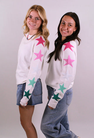 Two women wearing white sweaters with colorful star patterns on a plain background