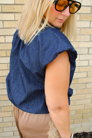 A woman wearing a dark blue denim-like short-sleeve top.  Back and sleeve detail of Breken dark wash denim bubble top, She is wearing large black-framed sunglasses with orange lenses. Available at Erin Rose Boutique