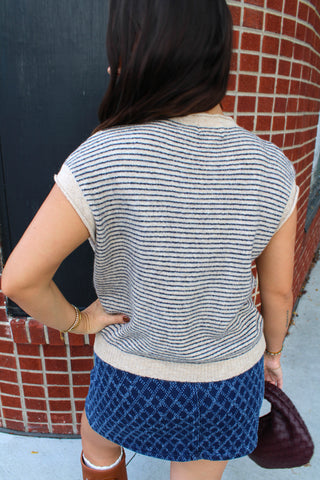 A woman viewed from behind wearing a cream and navy blue horizontally striped short-sleeve knit top and a blue patterned skirt, standing outdoors next to a brick wall.
