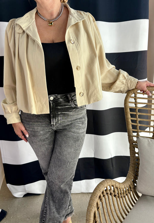 Person wearing a beige jacket and gray jeans in front of a black and white striped curtain.