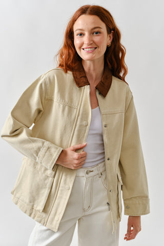 Front view of a woman wearing a beige utility barn jacket with brown corduroy collar, snap button closure, and front pockets.