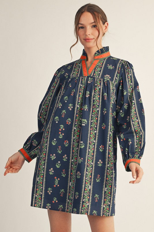 Woman wearing a navy floral tunic dress with ruffled neckline, long sleeves, and colorful trim details