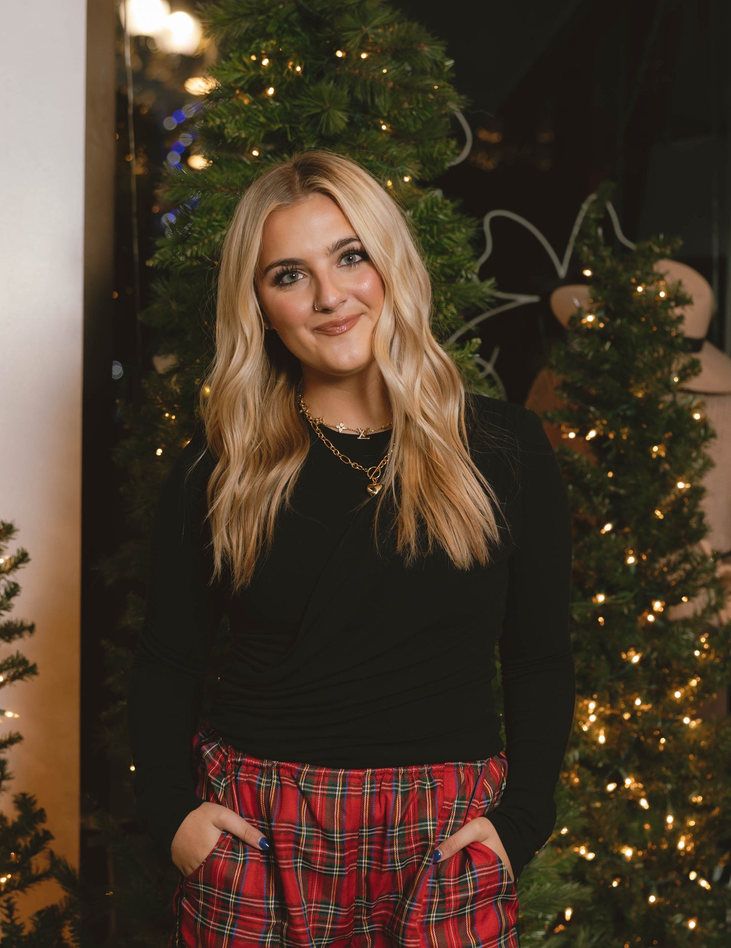 woman wearing black draped long sleeve top with red plaid pants in front of Christmas trees at Erin Rose Boutique