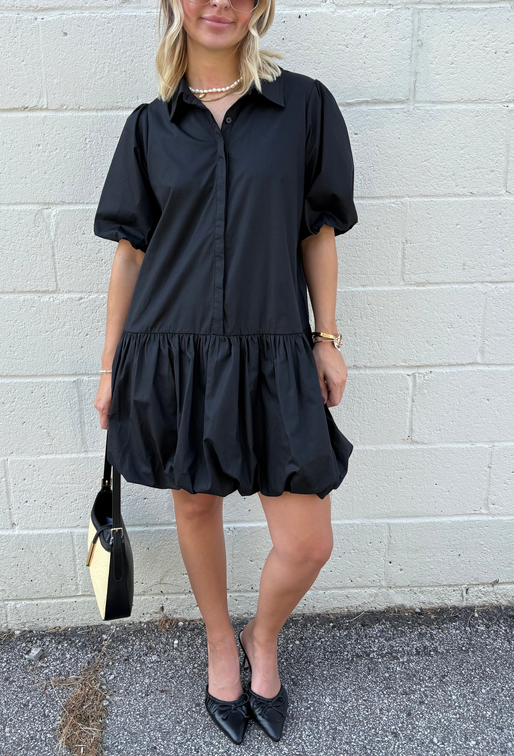 Woman wearing a black dress with puff sleeves against a gray brick wall.