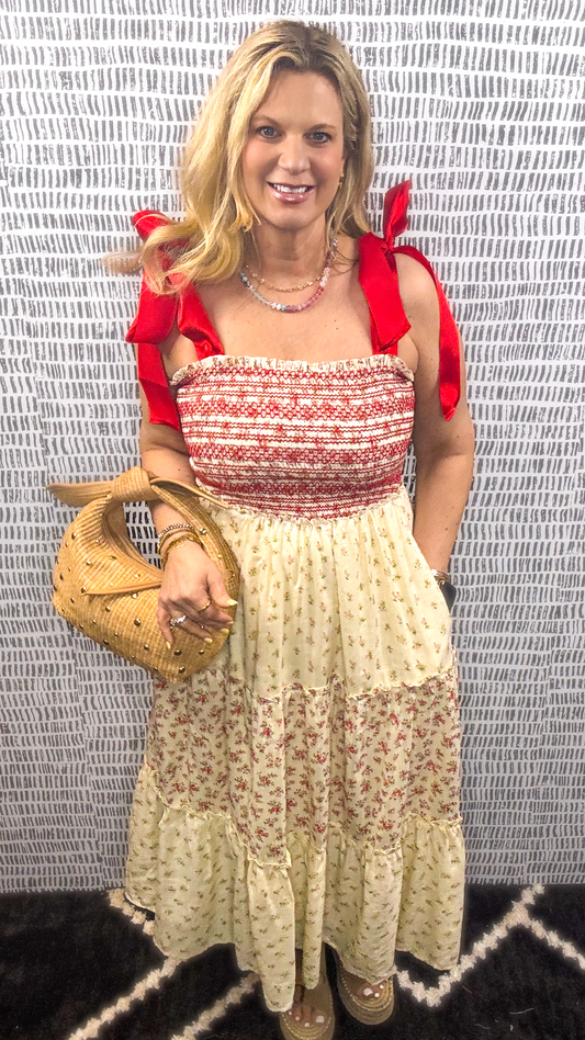 Woman wearing a red smocked midi dress with red tie straps and a tiered floral skirt, styled with a straw handbag and platform sandals