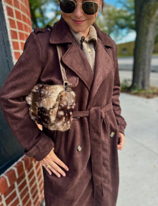 A woman wearing a long brown faux suede-like trench coat with a matching belt, styled with a brown and white patterned faux-fur shoulder bag and sunglasses, standing outdoors on a sidewalk next to a brick wall.