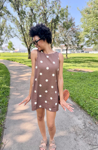 Woman in a brown polka dot shift dress standing on a sidewalk with trees
