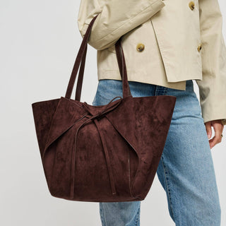 A dark brown suede tote bag with a large front bow detail, carried by a model wearing a beige jacket and blue jeans against a plain light gray background.