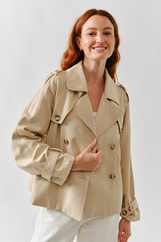 Woman wearing a beige cropped trench coat with double breasted buttons and belted sleeves.