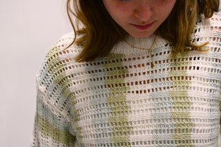 Close-up of a person wearing a loose open-knit sweater with a white base and soft sage green stripes, against a plain light background.  Women’s cream and sage striped knit sweater available at Erin Rose Boutique.