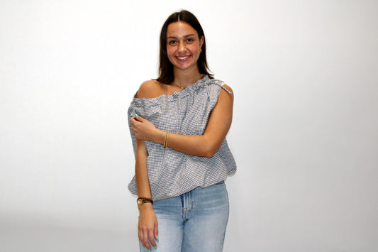Model wearing a blue and white gingham one-shoulder top with tie detail, styled with light wash jeans.