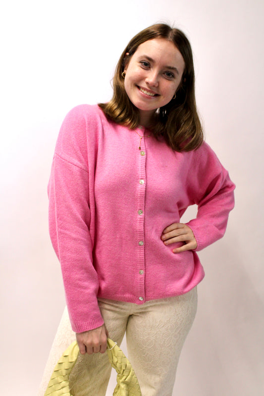 Model wearing a bright pink button-front knit cardigan styled with cream lace pants and holding a small yellow handbag.