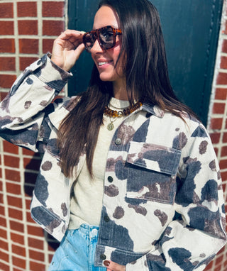 A woman wearing a distressed cow-print button-up shirt jacket over a cream knit top and light blue jeans, posing outdoors against a brick wall and dark green door.