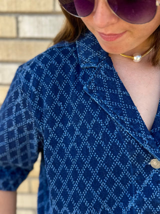 A close-up of a person wearing a blue short-sleeve collared top with a white geometric diamond pattern, styled with dark sunglasses and a pearl necklace against a light brick wall background.