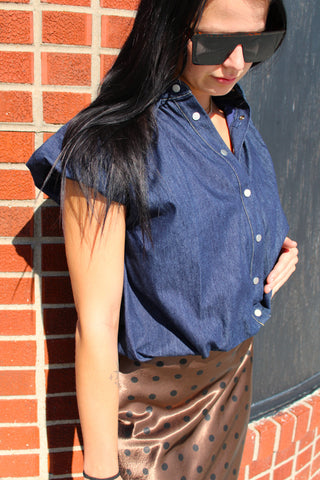 A woman wearing a short-sleeve blue denim button-up shirt and a brown satin skirt with black polka dots, standing outdoors against a red brick wall and a dark wall.