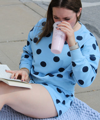 Woman in blue polka dot lounge set  drinking a pink drink and reading a book outdoors.