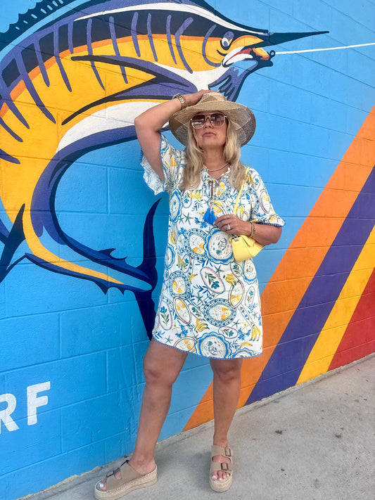 Woman wearing a lemon and tile print dress, straw hat, and platform sandals posing in front of a vibrant marlin mural with beachy summer vibes