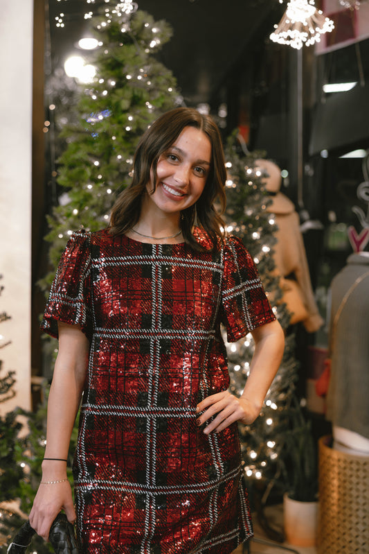 Woman in a red and black checkered dress standing in front of Christmas trees available at Erin Rose Boutique.