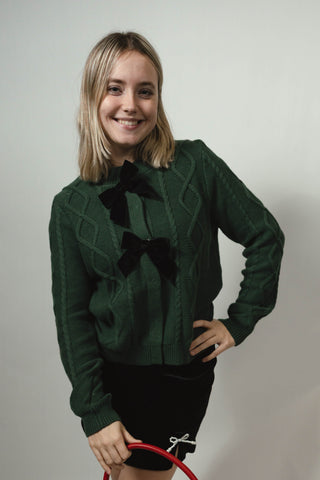 Woman wearing a green cardigan sweater with black bows and holding a red handbag on a white background