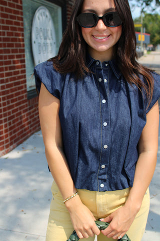 A woman wearing a dark blue chambray button-down shirt and light yellow pants, standing outdoors on a sidewalk next to a brick building.