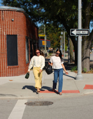 Two women walking on a city sidewalk next to a red brick building, one wearing a cream floral long-sleeve top and yellow skirt holding a green handbag, and the other wearing a white floral top and blue jeans holding a black handbag.