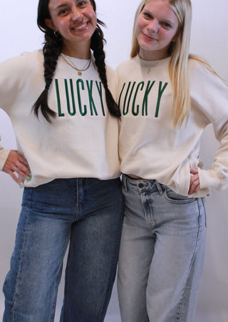 Two people wearing matching cream sweaters with 'LUCKY' printed on them, standing against a white background.