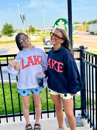 Two women smiling outside a coffee shop wearing oversized "LAKE" gingham graphic crewnecks paired with polka dot shorts and sunglasses.