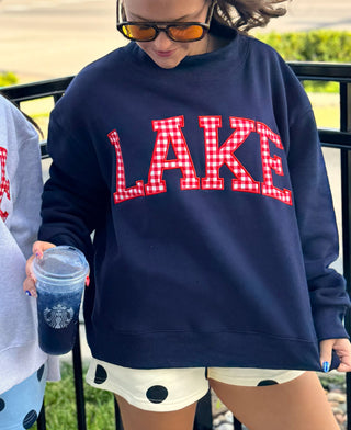 A person wearing an oversized navy sweatshirt with 'LAKE' in red-and-white gingham block letters, standing outdoors near a black railing and holding a Starbucks iced coffee.