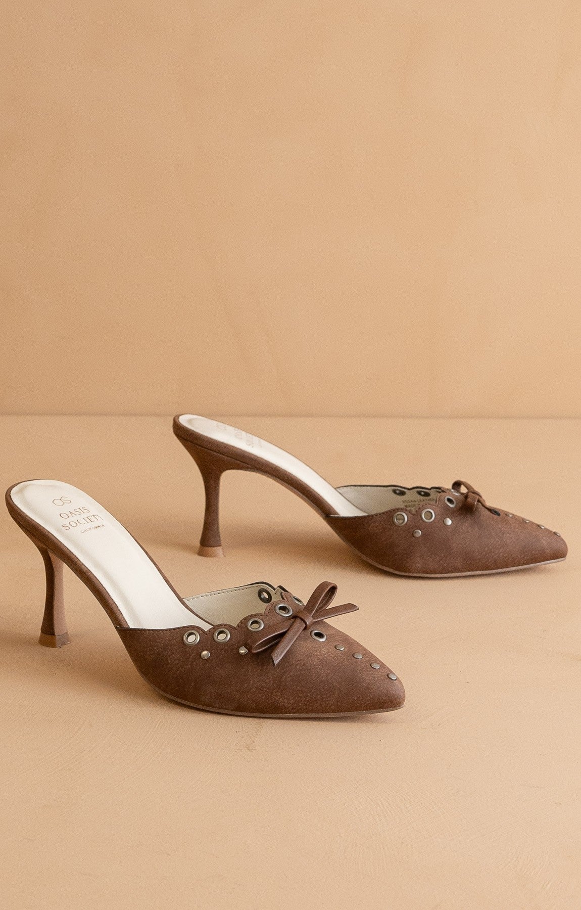 Pair of brown suede high-heeled shoes with decorative bows on a beige background