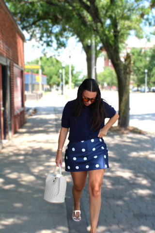 A woman walking on a sunny, tree-lined sidewalk wearing a navy blue short-sleeve top and a matching navy blue mini skirt with white polka dots. She is carrying a white woven tote bag.