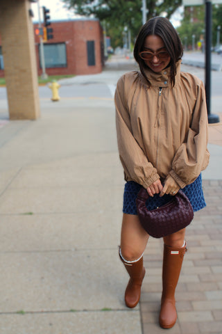 A woman stands on a city sidewalk wearing an Oversized funnel neck fall jacket for women blue patterned skirt, brown hunter knee-high boots, and holding a dark purple woven handbag.