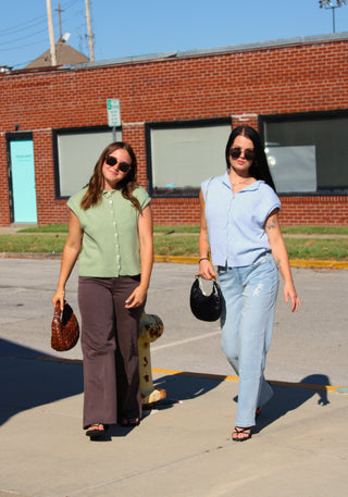 Two women walking outdoors on a sunny day in front of a brick building. The woman on the left wears a sage green short-sleeve button-up shirt and dark brown wide-leg pants, holding a brown handbag.
