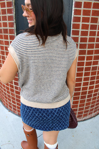 A woman viewed from behind wearing a beige and navy blue horizontally striped knit sweater vest, a blue patterned skirt, and brown boots, standing in front of a red brick tiled wall.
