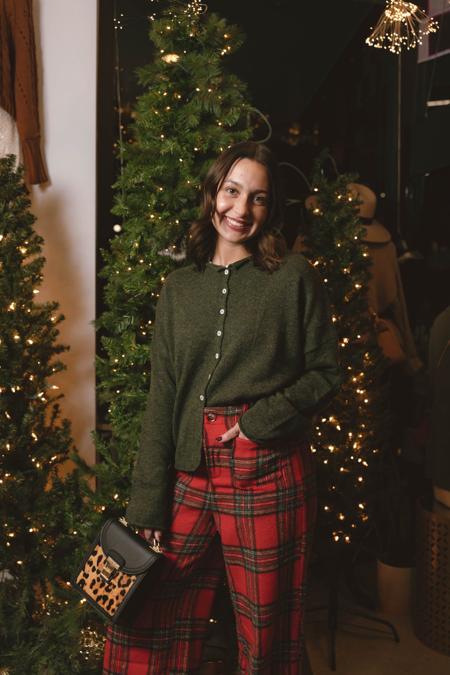Woman in a green cardigan and red plaid pants standing in front of Christmas trees with lights.