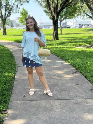 A woman stands on a paved path in a park wearing a light blue tunic top with a dark blue and white polka-dot skirt, carrying a beige handbag.