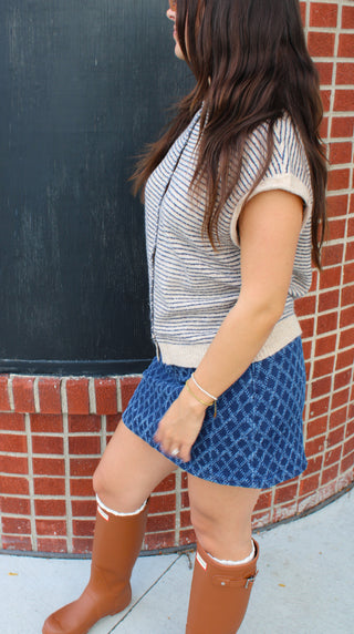 Side view of a woman wearing a striped, khaki, knit Piper top, a blue patterned mini skirt, and brown boots, standing in front of a brick wall.