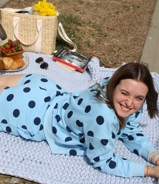 Woman in a polka dot lounge shorts with matching crew neck sweatshirt lying on a blanket with a picnic setup in the background.