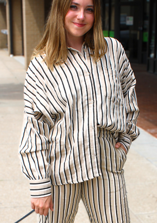A woman wearing a matching beige and black vertically striped lounge set, featuring a button-up shirt and pants, standing outdoors on a sidewalk.