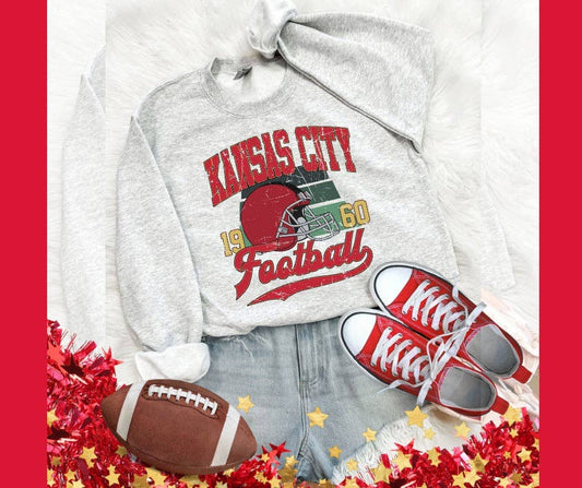 A vintage gray sweatshirt with 'KANSAS CITY 1960 FOOTBALL' text and a graphic design, accompanied by a pair of red and white sneakers and a football.