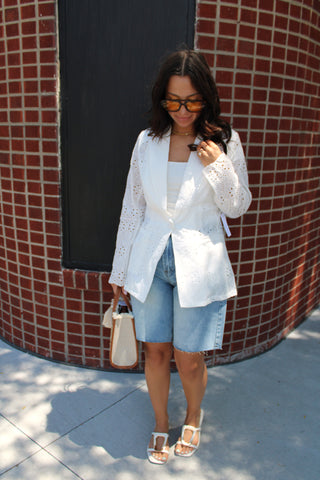 A woman stands outdoors in front of a red brick building, wearing a white eyelet blazer over a white top, light blue denim shorts, white heeled sandals, and holding a small white handbag.